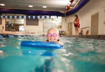 A girl enjoys swimming lessons in the indoor swimming pool and practices her kicks while using a kick board.