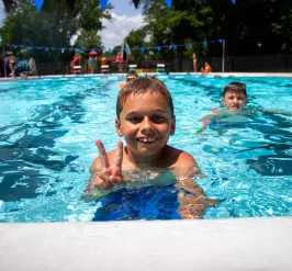 A smiling child shows a peace sign while swimming in the Kennett YMCA outdoor pool on a sunny day