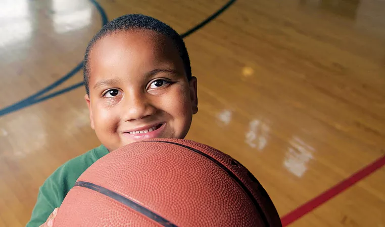 A young boy smiles at the camera while holding a basketball on the indoor basketball court.