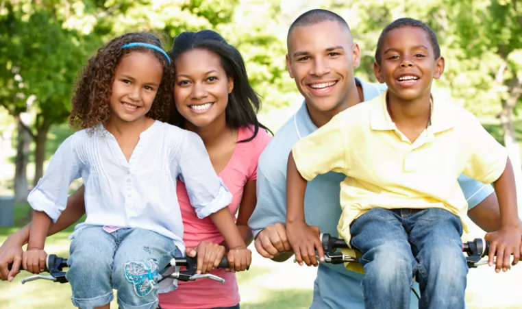A diverse YMCA family of four showing a mother, father, elementary-aged son and daughter pose with bicycles while smiling at the camera. 