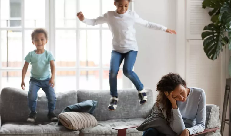 Two kids jump on the couch while a woman, their mom, sits with her head in her hands.