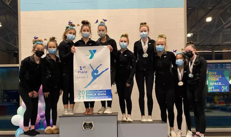 The YMCA Gymcats competitive gymnastics team gymnasts pose on the podium with their first place banner at a gymnastics competition. 