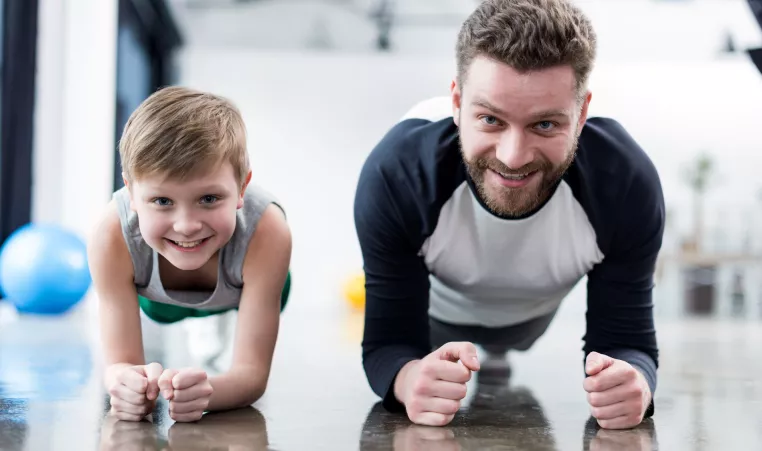 A father and son participate in a fitness class at the YMCA doing planks.