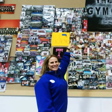 Jill Gambale, a gymnastics coach at the Oscar Lasko YMCA, poses next to a bulletin board showcasing many years of gymnastics team images