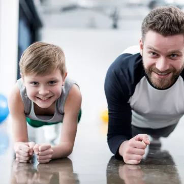 A father and son participate in a fitness class at the YMCA doing planks.